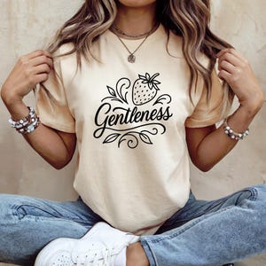May include: Cream-colored t-shirt featuring a black strawberry graphic with the word "Gentleness" in cursive script. The design includes leaves and swirls. The model is wearing blue jeans and white sneakers.