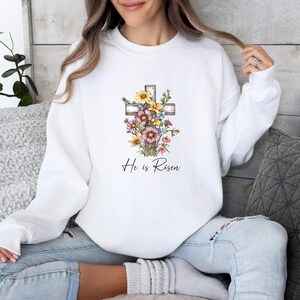 Pode incluir: Camisola branca com um design de cruz floral e o texto "He is Risen".
