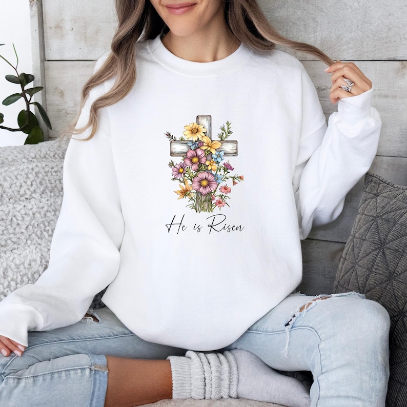 Pode incluir: Camisola branca com um design de cruz floral e o texto "He is Risen".