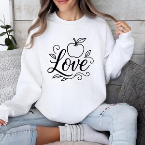 May include: White sweatshirt with a black apple and the word "Love" in a cursive font. The apple has leaves and the design includes decorative swirls. The sweatshirt is long-sleeved and has a crew neck.