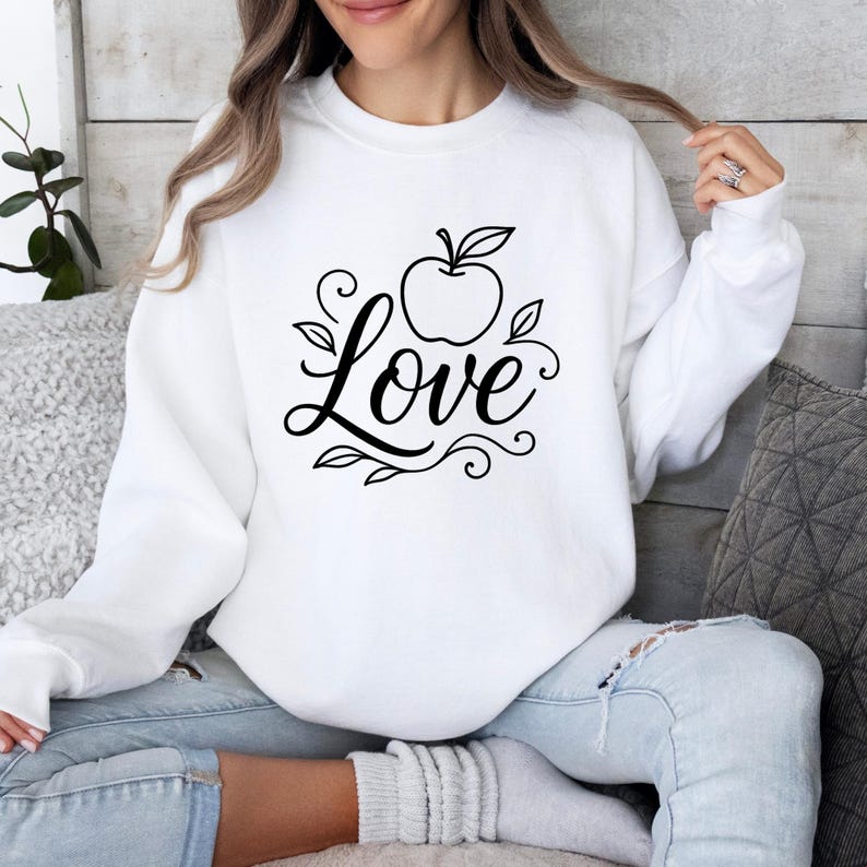 May include: White sweatshirt with a black apple and the word "Love" in a cursive font. The apple has leaves and the design includes decorative swirls. The sweatshirt is long-sleeved and has a crew neck.