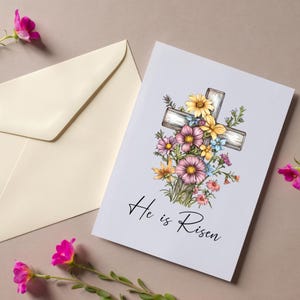 Pode incluir: Um cart&atilde;o branco com uma cruz de madeira decorada com flores coloridas. O texto "He is Risen" est&aacute; impresso abaixo da cruz.