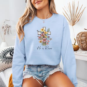 Pode incluir: Camisola azul claro com um design de cruz floral e o texto "He is Risen".