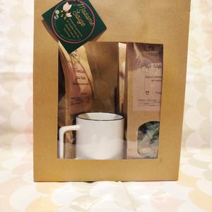 Puede incluir: Un juego de regalo en una bolsa de papel marrón con una ventana, que contiene una taza blanca con borde negro, paquetes de té y una etiqueta verde con el texto "Madame Sauge". La bolsa tiene asas de papel.