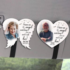 May include: Two memorial car visor ornaments. One is angel wing shaped with a photo of a woman and the text "I have an angel watching over me and I call her MOM 1960-2025". The other is heart-shaped with a photo of a man and the text "I have an angel watching over me and I call him DAD 1960-2025".