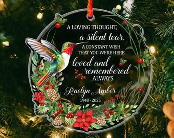 May include: A round clear acrylic ornament featuring a hummingbird, red poinsettias, pine cones, and holly berries. The text reads: A LOVING THOUGHT, a silent tear, A CONSTANT WISH THAT YOU WERE HERE loved and remembered ALWAYS Raelyn Amber 1948-2025.