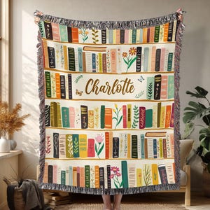 May include: A cream-colored throw blanket featuring a bookshelf design with colorful books, flowers, and the name "Charlotte" in a script font. The blanket has a fringed edge and is displayed indoors.
