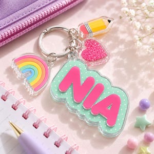 May include: A keychain featuring a mint green acrylic charm with white polka dots and the name NIA in bold pink letters. Attached are three smaller charms: a colorful rainbow, a yellow pencil, and a glittery pink heart on a silver ring.