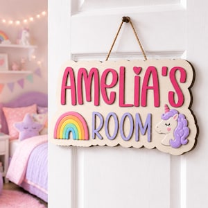 May include: A light-colored wooden door sign with a scalloped edge, hanging by a brown rope. The sign features the text "AMELIA'S ROOM" in pink and purple, a colorful rainbow graphic, and a purple unicorn head with a pink horn and closed eyes.