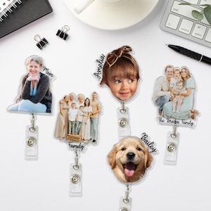 May include: A collection of personalized photo ID badge reels. Each reel features a different image, including a family portrait, a child's face, a dog, and a woman. The reels have a clear plastic body and a retractable cord.