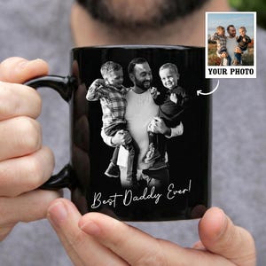 May include: A white ceramic mug with a black handle and the text "But First, Coffee" in a bold, black font. The mug is set against a blurred background, suggesting a cozy setting.