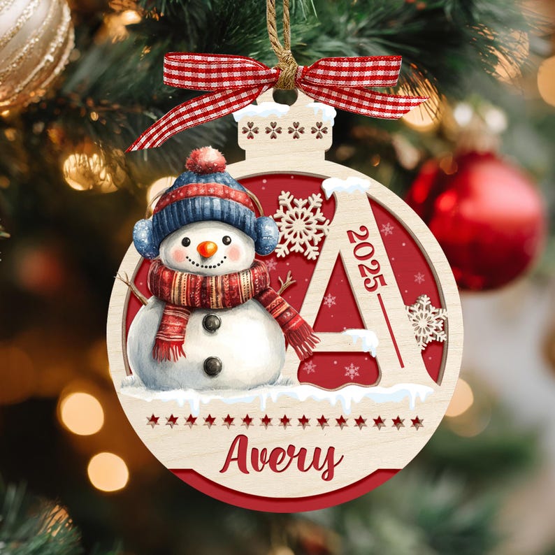 May include: A round wooden Christmas ornament featuring a snowman wearing a blue hat and red scarf, a large letter A with the year 2025, and the name Avery printed below. The ornament is topped with a red and white checkered fabric ribbon bow.