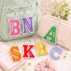 May include: A collection of seven acrylic letter keychains featuring various colors and patterns, each personalized with a name. The letters B, N, A, S, K, H, and C are displayed with silver metal rings, showcasing floral, solid, and gingham designs.