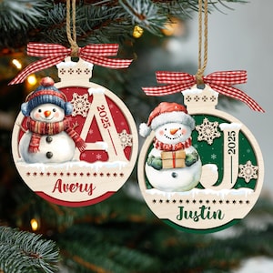 May include: Two round wooden Christmas ornaments with red and green backgrounds, each featuring a snowman, a snowflake, the year 2025, and a name. The left ornament says Avery and the right says Justin. Both have red gingham bows and twine.