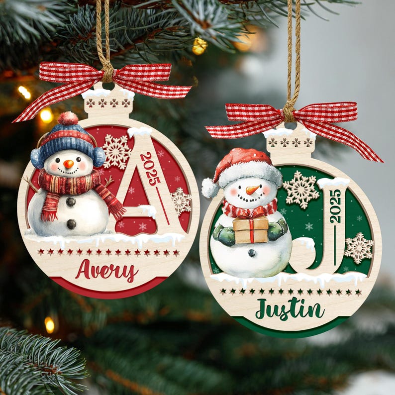 May include: Two round wooden Christmas ornaments with red and green backgrounds, each featuring a snowman, a snowflake, the year 2025, and a name. The left ornament says Avery and the right says Justin. Both have red gingham bows and twine.