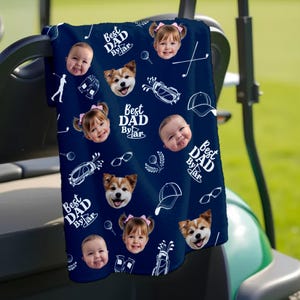 Puede incluir: Toalla de golf azul marino con un patr&oacute;n repetido de caras de beb&eacute;s, caras de perros, gr&aacute;ficos de golf y el texto "Best Dad By Far" en blanco. La toalla est&aacute; sobre un carrito de golf.