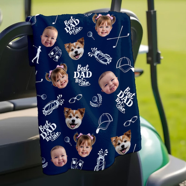 Puede incluir: Toalla de golf azul marino con un patr&oacute;n repetido de caras de beb&eacute;s, caras de perros, gr&aacute;ficos de golf y el texto "Best Dad By Far" en blanco. La toalla est&aacute; sobre un carrito de golf.