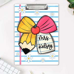 May include: A white clipboard with a whimsical design featuring a yellow pencil, a pink bow, and a red apple. The clipboard has blue stripes, daisy accents, and the text "Mrs. Kelsey".
