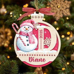 May include: A round wooden Christmas ornament featuring a snowman wearing a pink hat and scarf, holding a candy cane. The design includes a large letter D, the year 2025, and the name Diane in script. A red and white gingham ribbon is tied at the top.