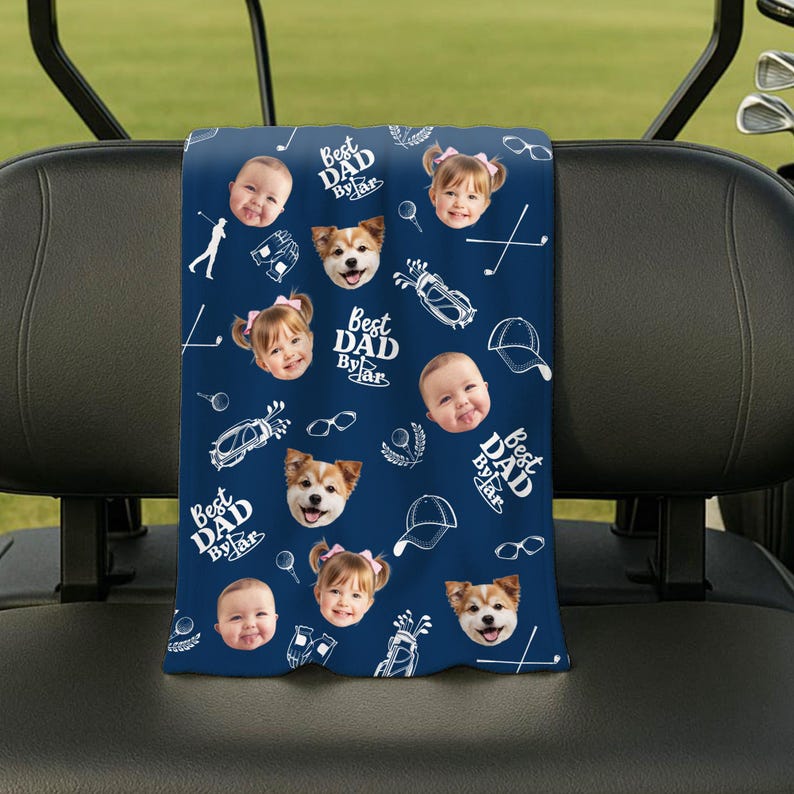 Puede incluir: Una toalla de golf azul marino con un patr&oacute;n repetido de caras de beb&eacute;s y perros, ilustraciones tem&aacute;ticas de golf y el texto "Best Dad By Far" en blanco. La toalla est&aacute; colgada en un carrito de golf.