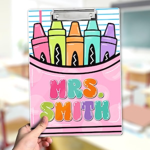 May include: A pink clipboard featuring a colorful crayon design and the text "MRS. SMITH." The design includes pink, green, orange, blue, and purple crayons. The clipboard has a silver metal clip at the top.