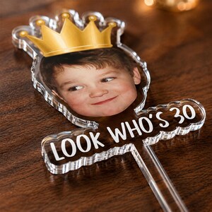 Puede incluir: Un adorno para tarta de acr&iacute;lico transparente con una fotograf&iacute;a impresa de un ni&ntilde;o con una ilustraci&oacute;n de corona dorada. El adorno incluye el texto blanco "LOOK WHO'S 30" y tiene un tallo transparente para insertar en la tarta.
