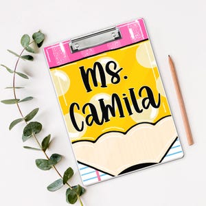 May include: A clipboard with a yellow pencil design and the text "Ms. Camila" in black. The top of the clipboard is pink with a silver clip. A wooden pencil and green leaves are next to the clipboard.