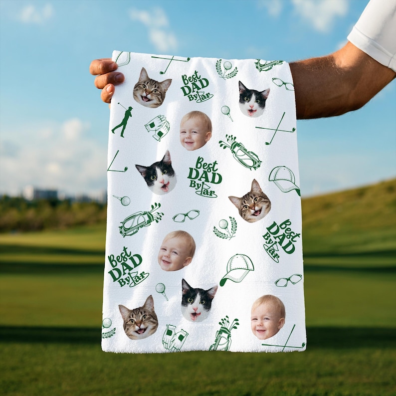 Puede incluir: Toalla de golf blanca con un patr&oacute;n repetido de caras de gatos, caras de beb&eacute;s, gr&aacute;ficos tem&aacute;ticos de golf y el texto "Best Dad By Far" en verde. La toalla se muestra contra un campo de golf verde y un cielo azul.