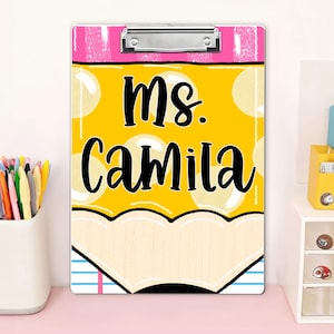 May include: A clipboard with a pencil design, featuring the name "Ms. Camila" in black lettering. The clipboard has a yellow background with white polka dots, a pink top, and a silver clip. The bottom of the clipboard has a light wood-like design.