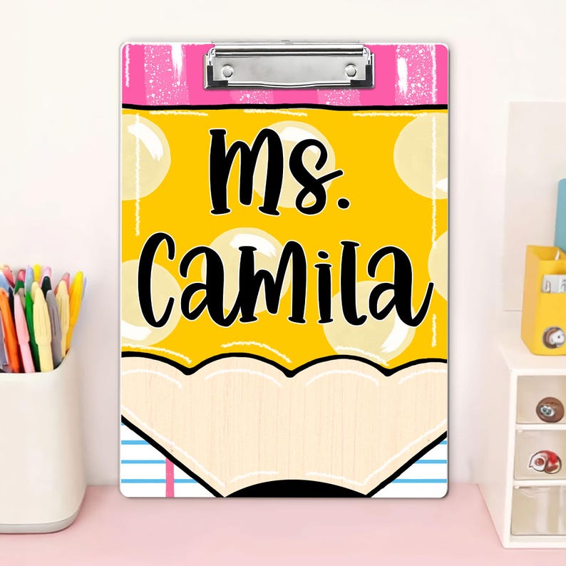 May include: A clipboard with a pencil design, featuring the name "Ms. Camila" in black lettering. The clipboard has a yellow background with white polka dots, a pink top, and a silver clip. The bottom of the clipboard has a light wood-like design.