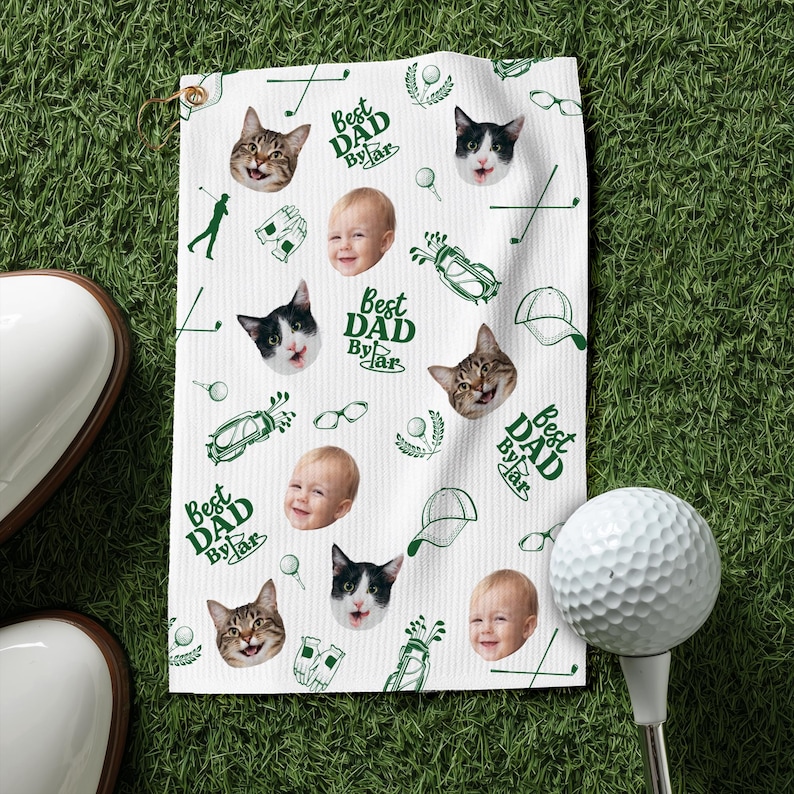Puede incluir: Toalla de golf blanca con un patr&oacute;n repetido de caras de gatos, caras de beb&eacute;s, ilustraciones tem&aacute;ticas de golf y el texto "Best Dad By Far" en verde. Una pelota de golf y un zapato de golf est&aacute;n en primer plano.