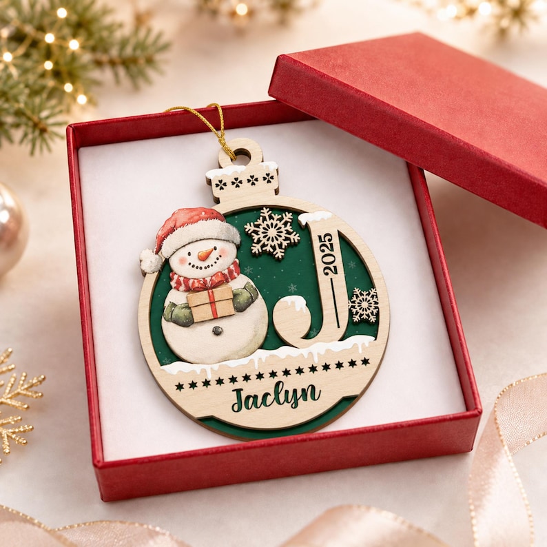 May include: A round wooden Christmas ornament featuring a snowman wearing a red hat and scarf, holding a gift. The ornament includes a cutout letter J, the year 2025, and the name Jaclyn printed in black. It is displayed inside a red gift box.