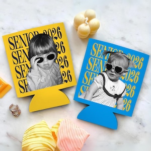 Puede incluir: Dos aislantes para bebidas, uno amarillo y otro azul, cada uno con una foto en blanco y negro de un ni&ntilde;o con gafas de sol. El texto "SENIOR 2026" est&aacute; impreso en amarillo en el aislante amarillo y en blanco en el azul. Tambi&eacute;n se ven una vela y accesorios.