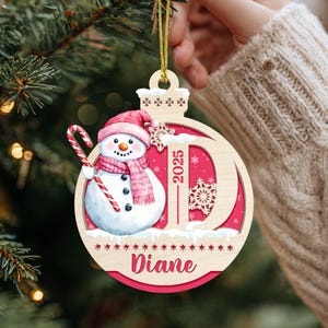 May include: A round wooden Christmas ornament featuring a snowman wearing a pink hat and scarf, holding a candy cane. The design includes a large letter D, the year 2025, and the name Diane in pink script, all set against a vibrant pink background.
