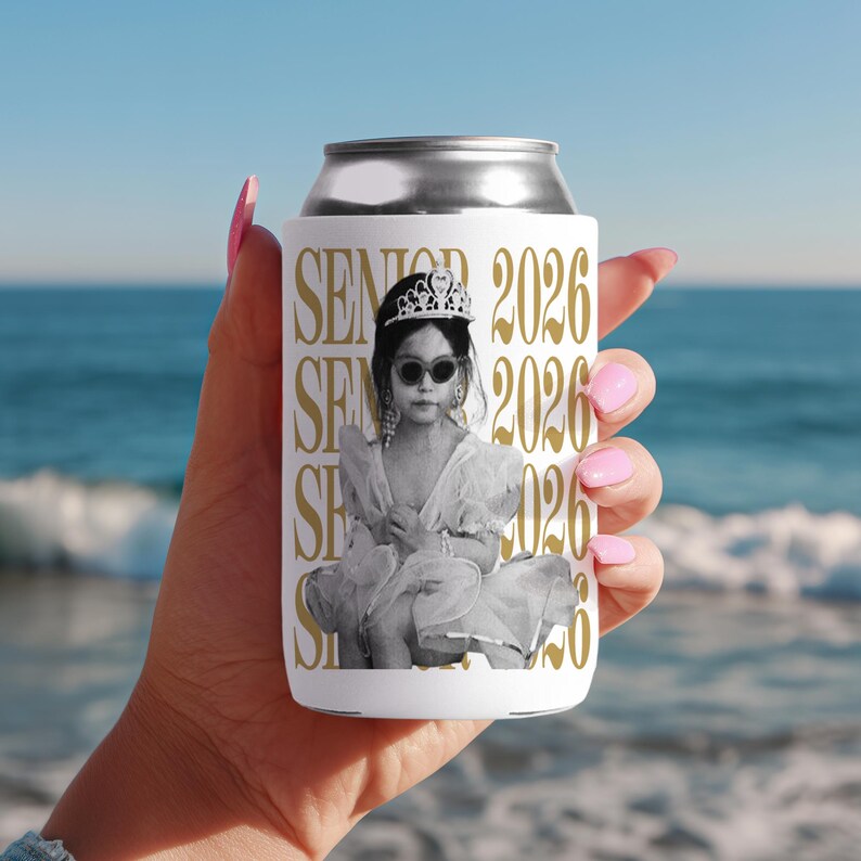 Puede incluir: Enfriador de latas blanco con un dise&ntilde;o dorado con las palabras "SENIOR 2026" y una imagen en blanco y negro de una ni&ntilde;a con gafas de sol y una tiara. El enfriador de latas est&aacute; sostenido por una persona con esmalte de u&ntilde;as rosa.