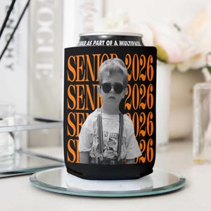 Puede incluir: Enfriador de latas negro con el texto "SENIOR 2026" en naranja, con una foto en blanco y negro de un ni&ntilde;o con gafas de sol. El enfriador est&aacute; sobre una superficie espejada.