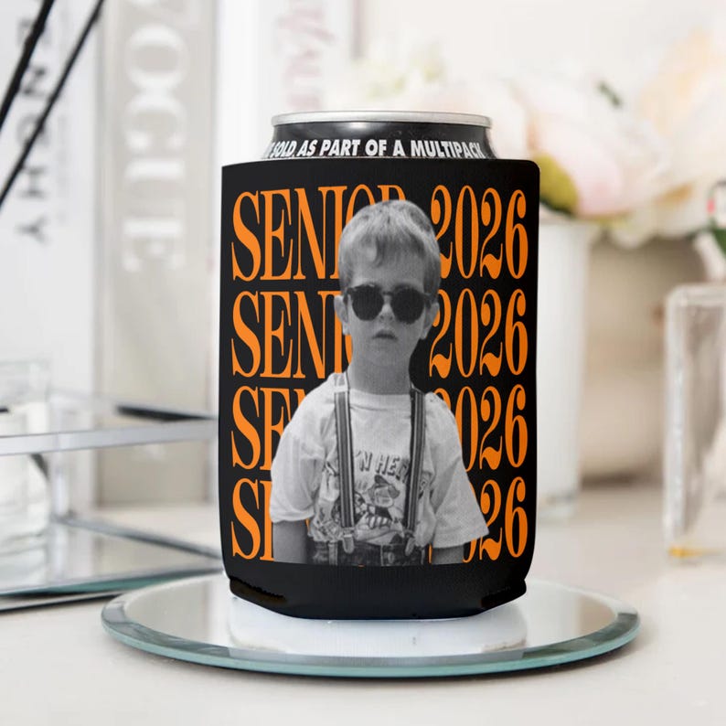 Puede incluir: Enfriador de latas negro con el texto "SENIOR 2026" en naranja, con una foto en blanco y negro de un ni&ntilde;o con gafas de sol. El enfriador est&aacute; sobre una superficie espejada.
