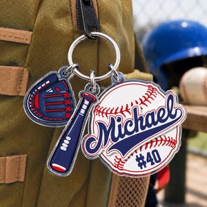 May include: A baseball-themed keychain featuring three acrylic charms attached to a silver metal ring. The charms include a navy blue baseball glove, a navy blue baseball bat, and a white baseball with red stitching, the name Michael, and #40.