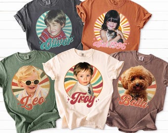 May include: Five vintage-style t-shirts in various colors, each featuring a circular portrait. The portraits include a child, a girl with a cat, a child with sunglasses, a child, and a dog. Each shirt has a name or year.