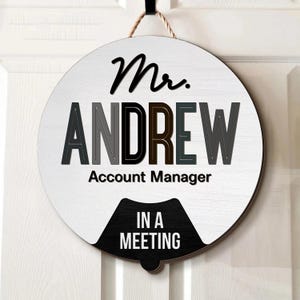 May include: A round, white wooden sign with the name "ANDREW" in gray, black, and brown block letters. The sign reads "Mr. ANDREW Account Manager IN A MEETING" in black text. A jute rope is used for hanging.