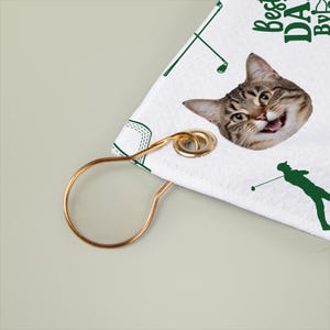 Puede incluir: Toalla de golf blanca con la cara de un gato y gr&aacute;ficos verdes con tem&aacute;tica de golf. La toalla tiene el texto "Best DA..." y un lazo de metal dorado para colgar. El gato tiene una expresi&oacute;n feliz.