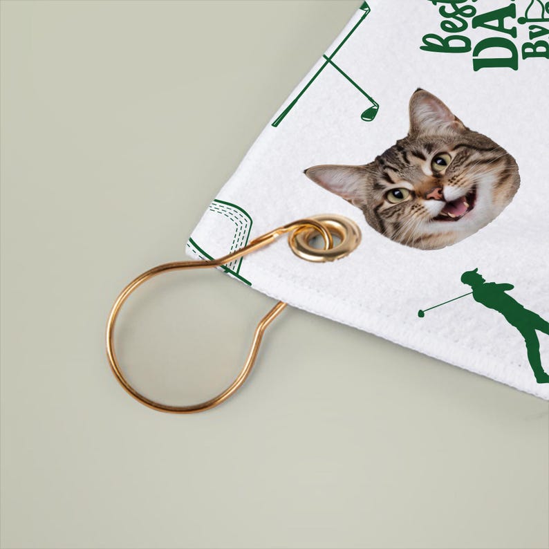 Puede incluir: Toalla de golf blanca con la cara de un gato y gr&aacute;ficos verdes con tem&aacute;tica de golf. La toalla tiene el texto "Best DA..." y un lazo de metal dorado para colgar. El gato tiene una expresi&oacute;n feliz.