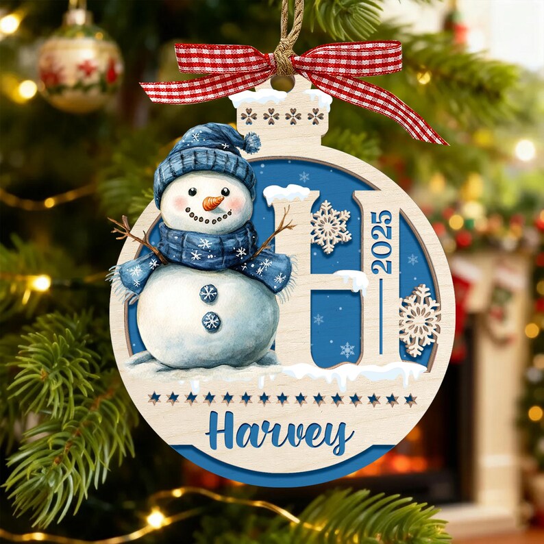 May include: A round wooden Christmas ornament featuring a snowman wearing a blue hat and scarf, a large letter H, the year 2025, and the name Harvey. The ornament is decorated with white snowflakes and topped with a red and white gingham ribbon.