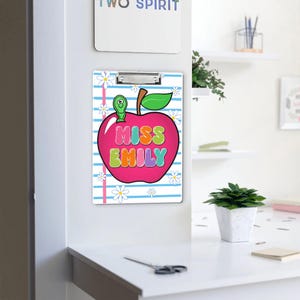 May include: A clipboard with a pink apple design, the apple has the text "MISS EMILY" in colorful bubble letters. The background has blue stripes and white daisy flowers. A green worm is on top of the apple.