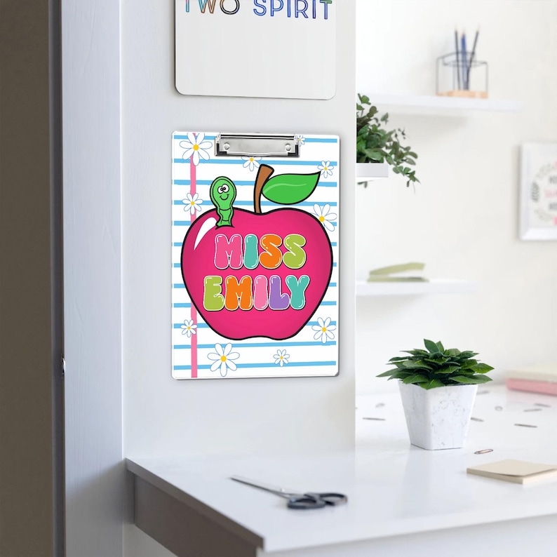 May include: A clipboard with a pink apple design, the apple has the text "MISS EMILY" in colorful bubble letters. The background has blue stripes and white daisy flowers. A green worm is on top of the apple.