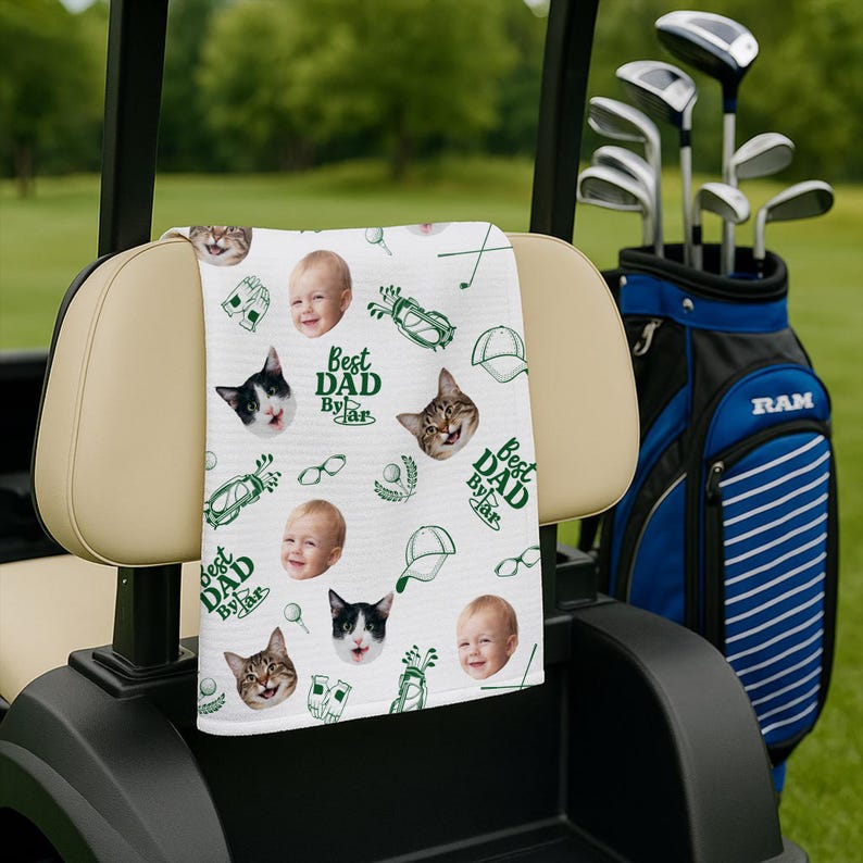 Puede incluir: Una toalla de golf blanca con un patr&oacute;n repetido de caras de beb&eacute;s, caras de gatos, ilustraciones tem&aacute;ticas de golf y el texto "Best Dad By Far" en verde. La toalla est&aacute; sobre el respaldo de un asiento de carrito de golf, con palos de golf en una bolsa azul y negra al fondo.