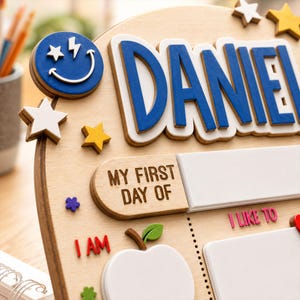 May include: A wooden sign featuring the name DANIEL in large blue letters, decorated with a blue smiley face, yellow and white stars, and a white apple shape. The board includes text that reads MY FIRST DAY OF, I AM, and I LIKE TO.