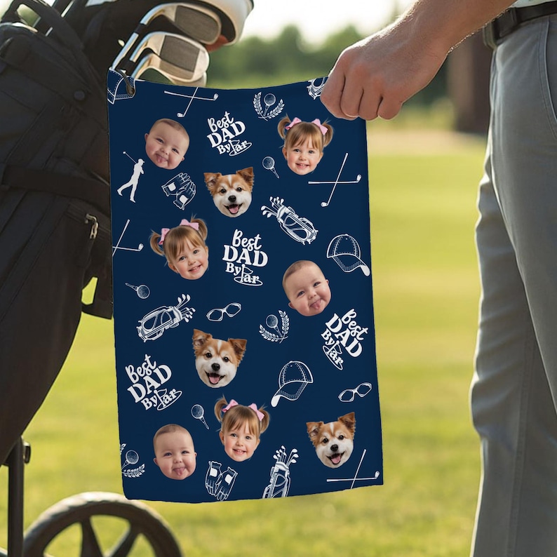 Puede incluir: Toalla de golf azul marino con un patr&oacute;n repetido de caras de beb&eacute;s, caras de perros, iconos de golf y el texto "Best Dad By Far" en blanco. La toalla se sostiene en un campo de golf.