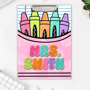 May include: A pink clipboard with a colorful crayon design. The design includes pink, green, orange, blue, and purple crayons in a pink pouch. The text "MRS. SMITH" is displayed in bubble letters.