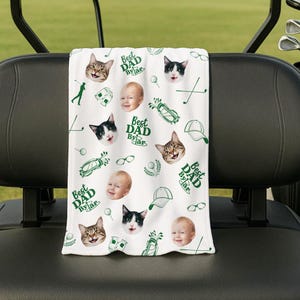 Puede incluir: Toalla de golf blanca con un patr&oacute;n repetido de caras de gatos, caras de beb&eacute;s, gr&aacute;ficos de golf y el texto "Best Dad By Far" en verde. La toalla est&aacute; colocada sobre el respaldo de un asiento de carrito de golf.